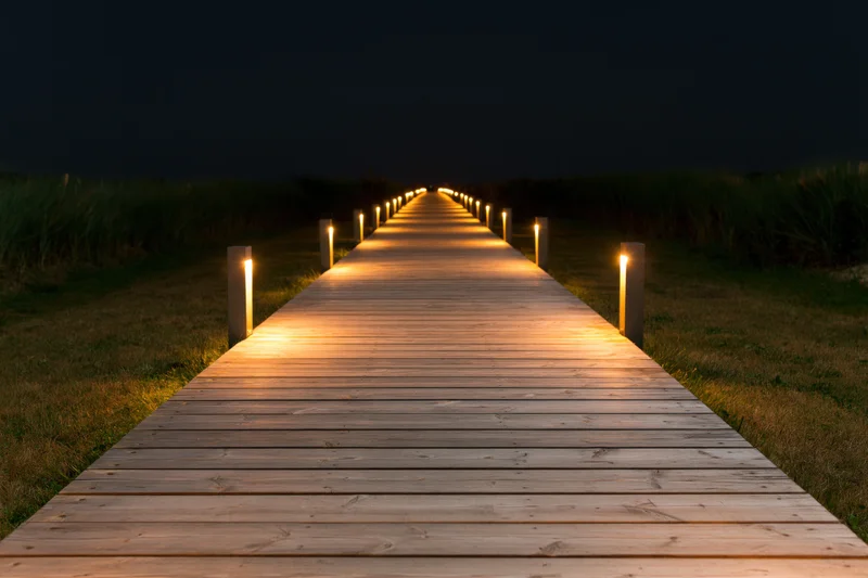 Wooden plank walkway illuminated by lanterns at night leading through grassy dunes