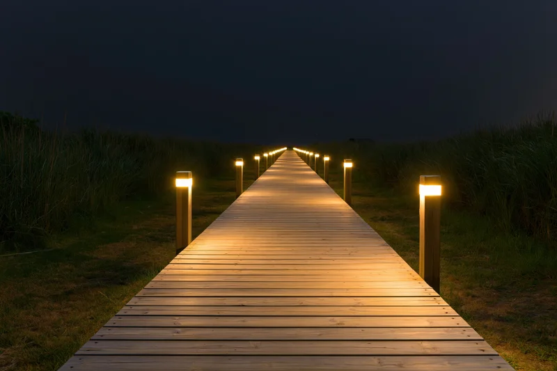 Wooden plank walkway illuminated by warm lights at night leading through grassy dunes toward the beach