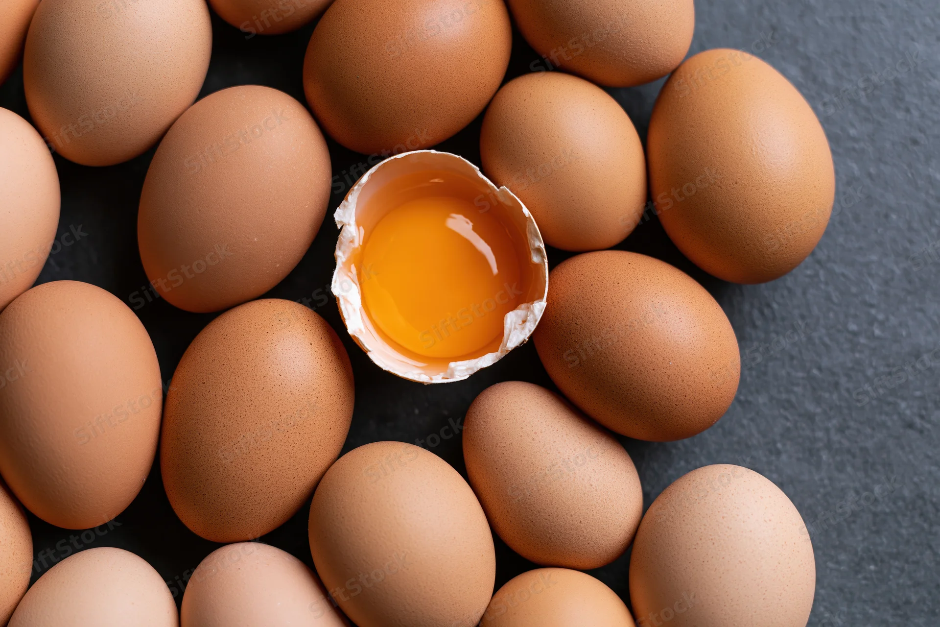 Brown chicken eggs arranged around one cracked egg with visible yolk on dark surface
