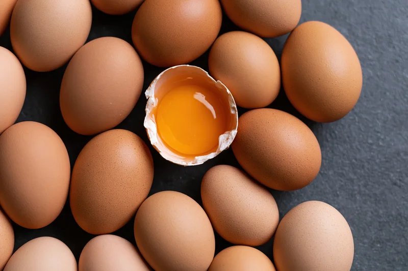 Brown chicken eggs arranged around one cracked egg with visible yolk on dark surface