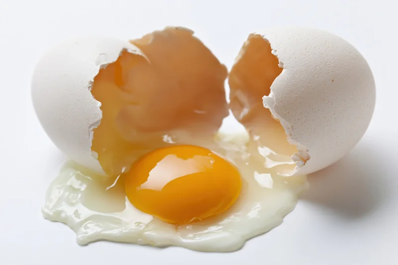 Cracked raw egg split open on pure white surface with yolk and white spilled out