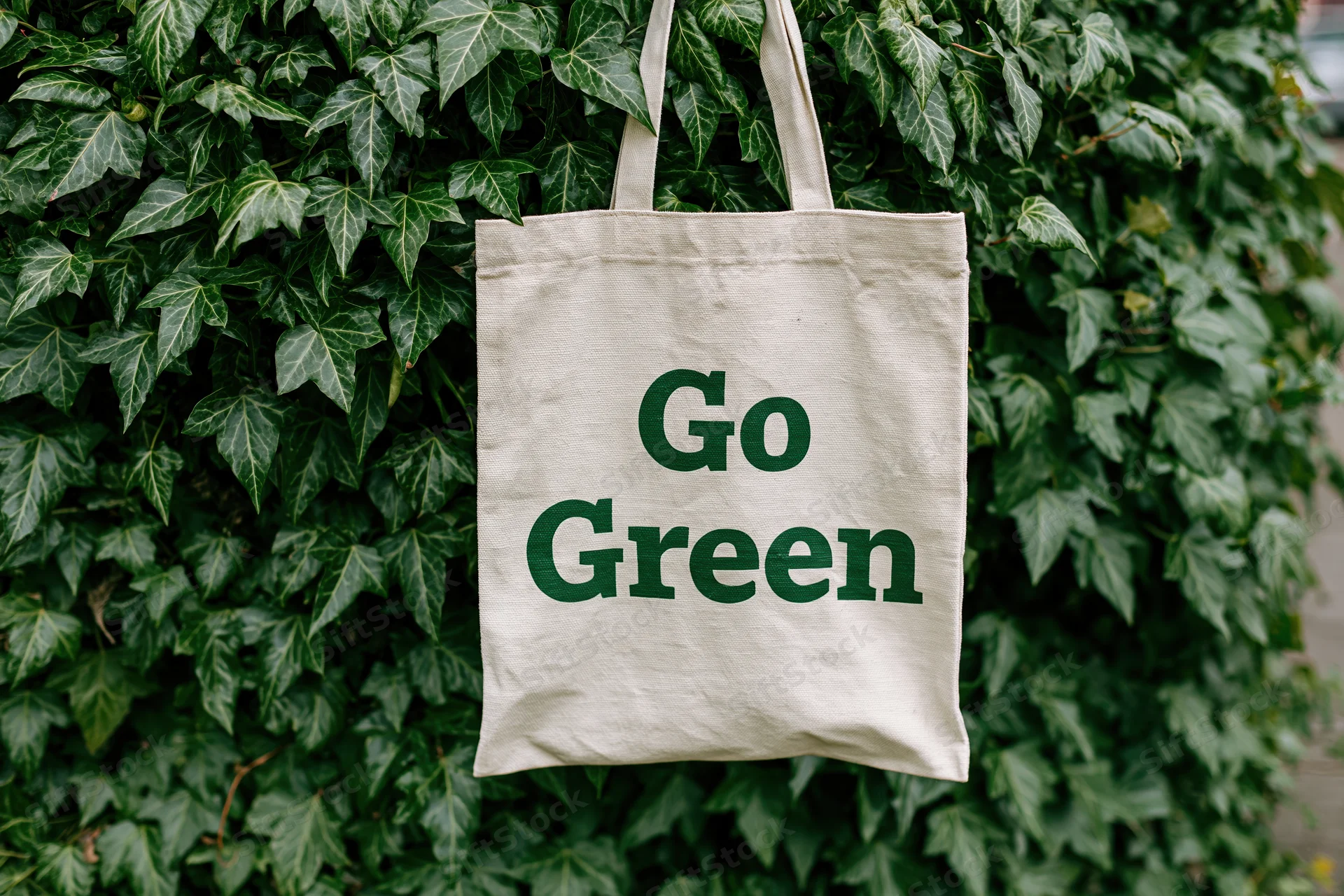 Cream canvas tote bag with bold dark green text reading Go Green hanging on a leafy ivy-covered wall