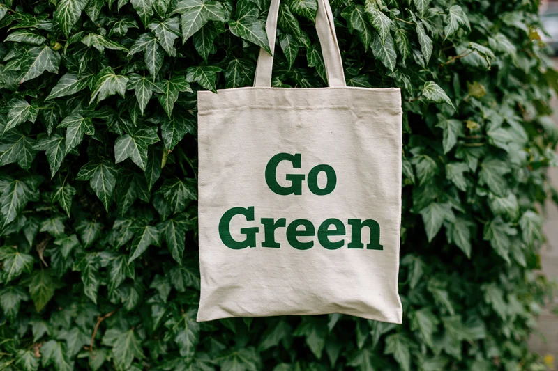 Cream canvas tote bag with bold dark green text reading Go Green hanging on a leafy ivy-covered wall