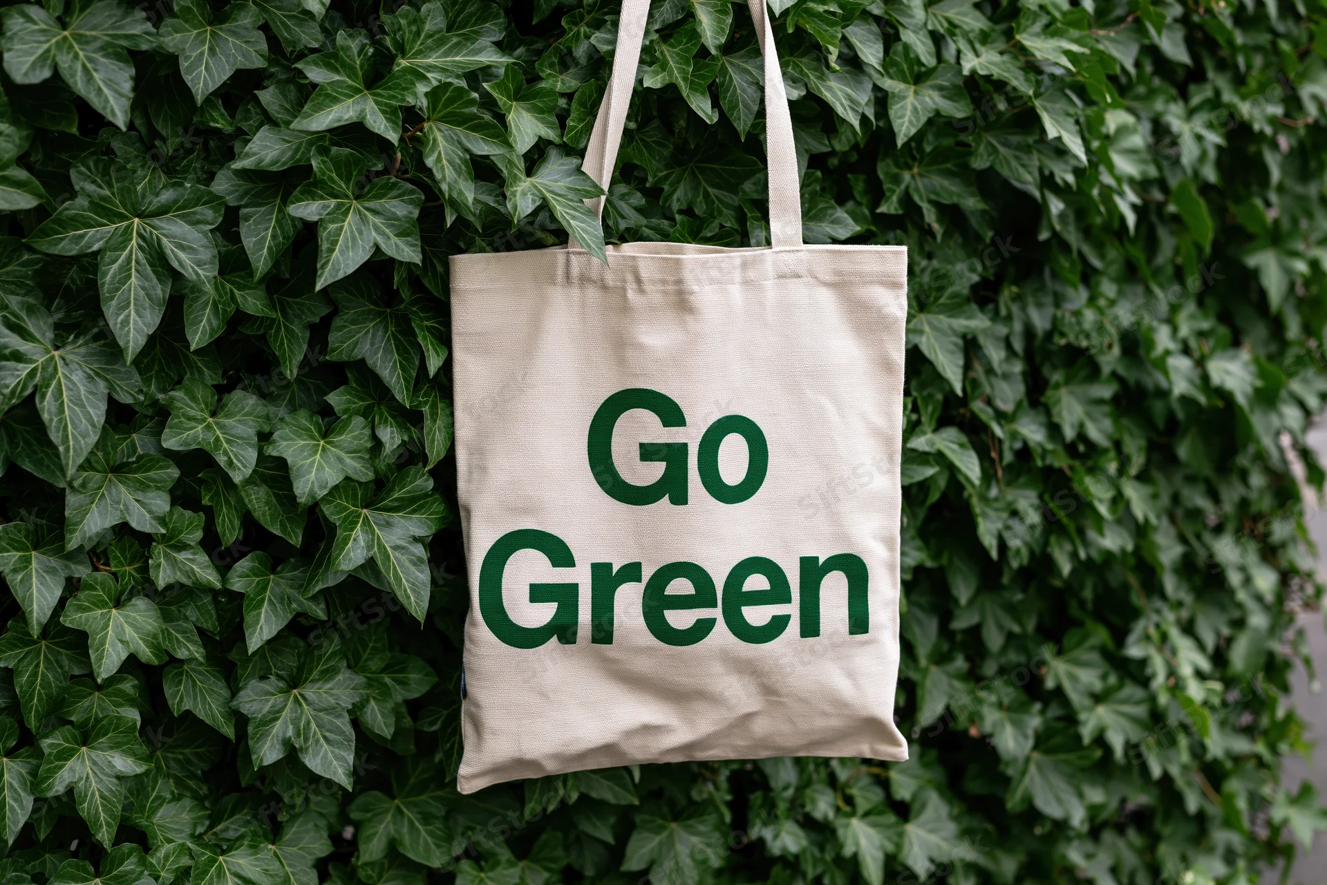 Cream canvas tote bag with bold dark green text reading Go Green hanging on a wall covered in green ivy foliage