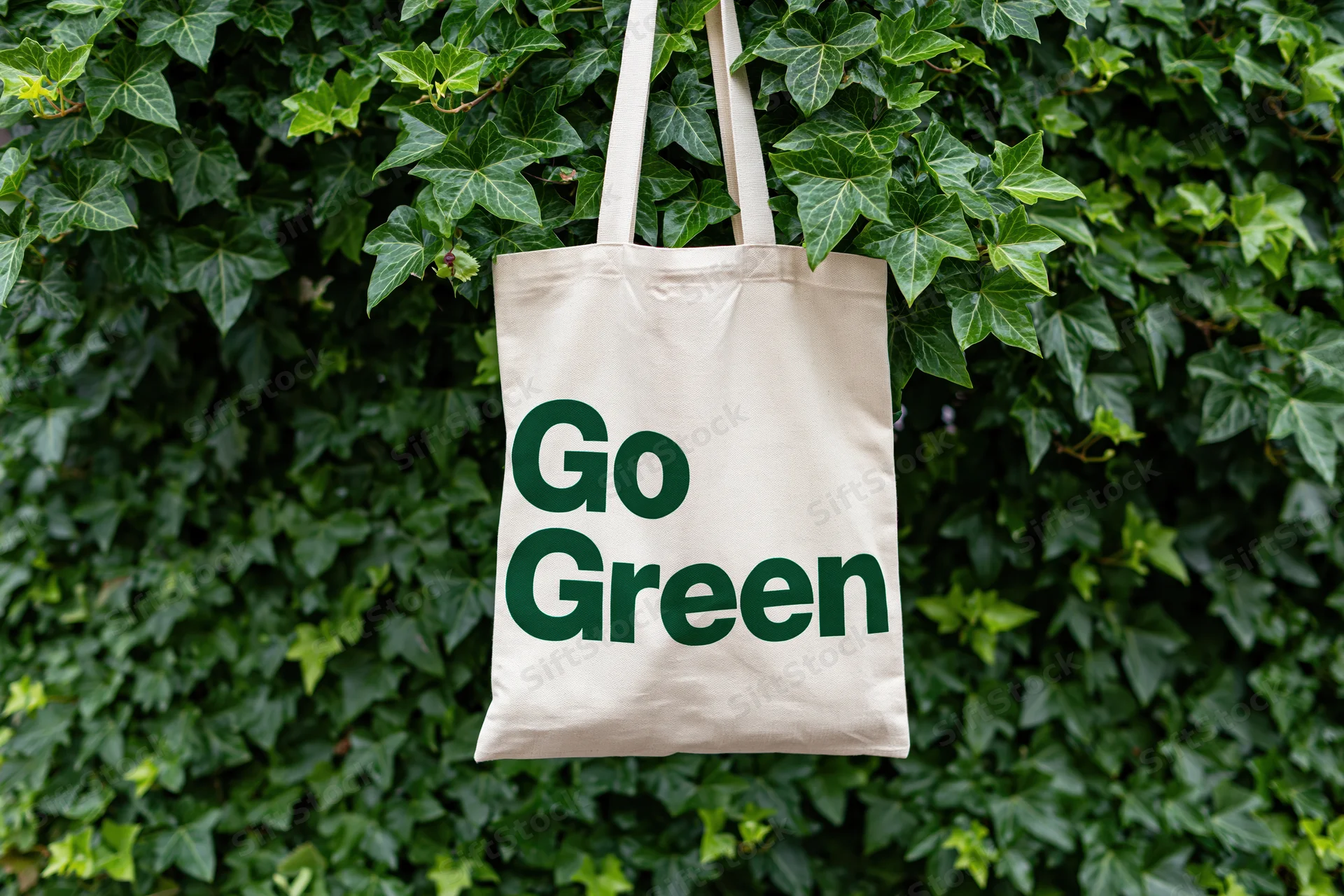 Cream canvas tote bag with bold dark green text reading Go Green hanging on ivy-covered wall
