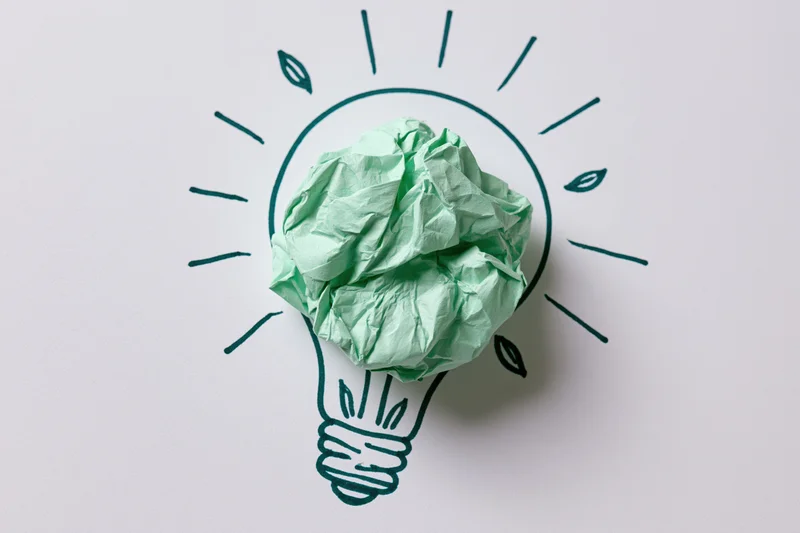 Crumpled green paper ball placed inside a hand-drawn lightbulb outline on white surface