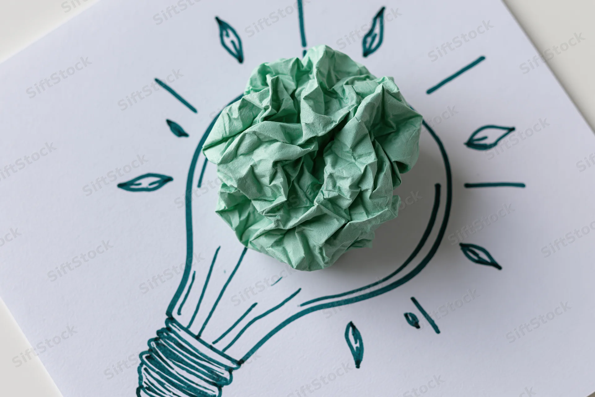Crumpled green paper ball placed on hand-drawn lightbulb sketch on white paper
