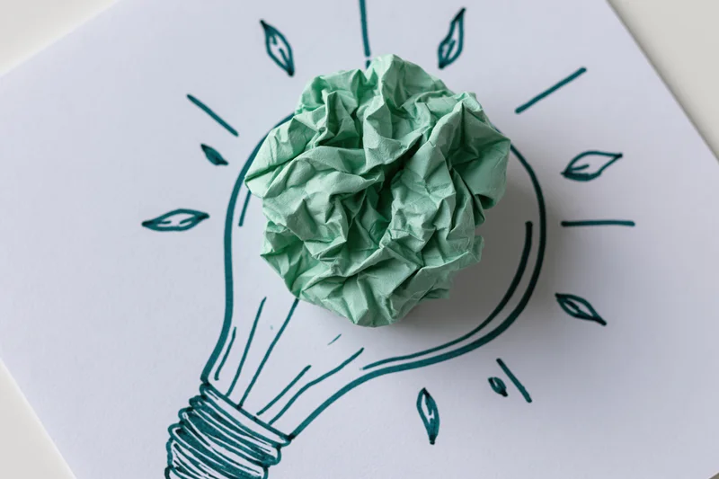 Crumpled green paper ball placed on hand-drawn lightbulb sketch on white paper