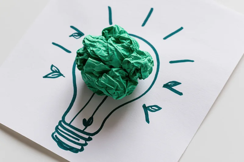 Crumpled green paper ball placed on a hand-drawn lightbulb sketch on white paper