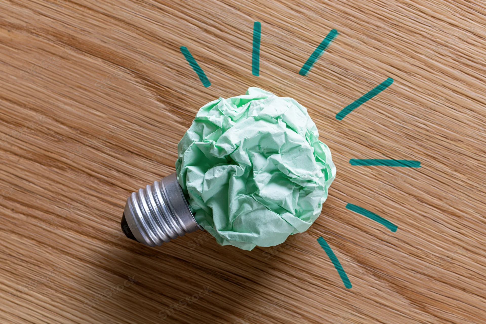 Crumpled green paper ball attached to silver lightbulb base on wooden surface with drawn light rays