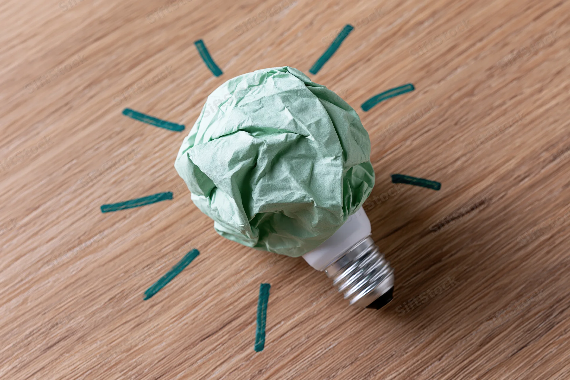 Crumpled green paper ball resting on a silver light bulb base on wooden surface with drawn green rays around it