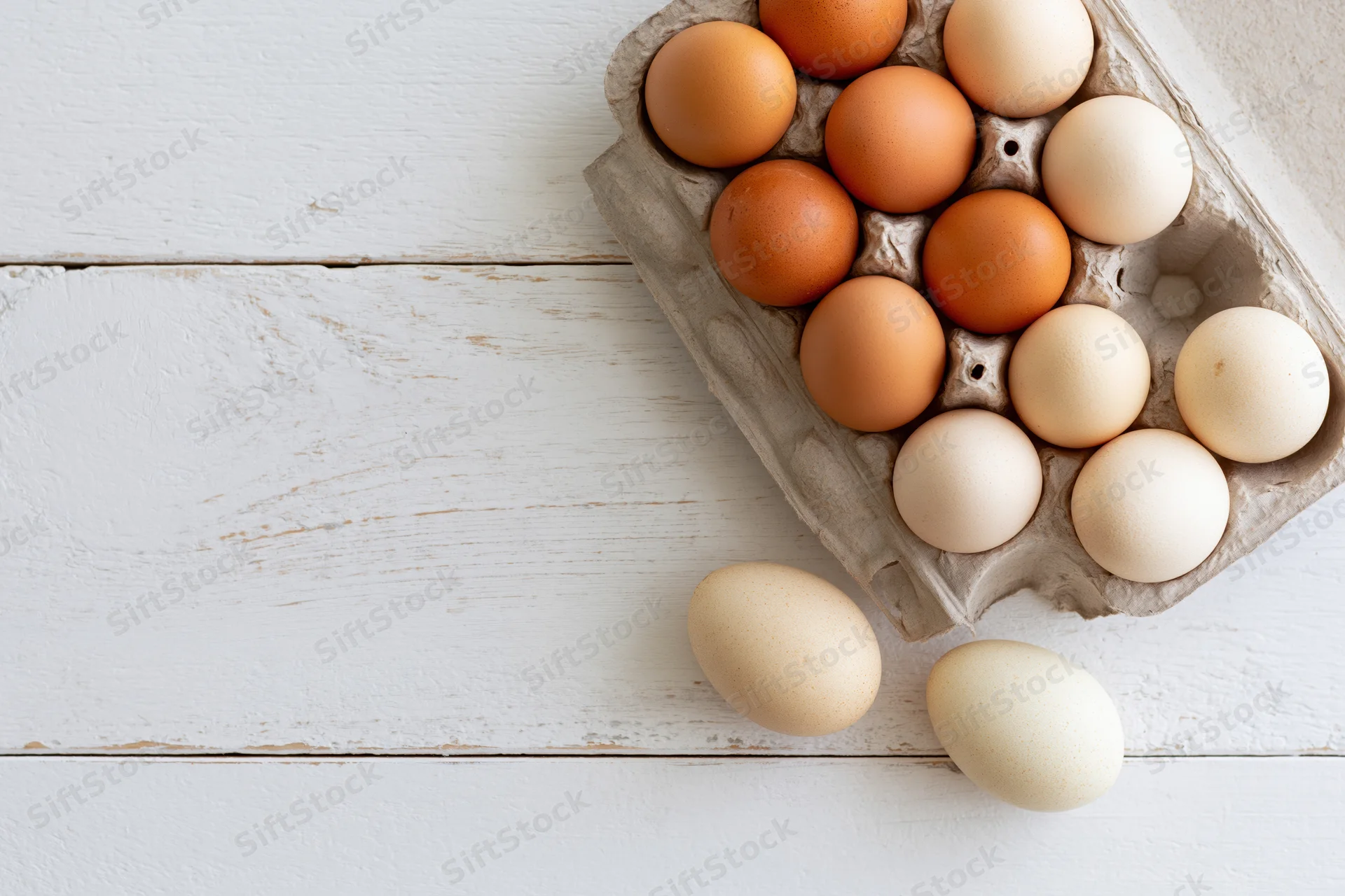 Farm-fresh chicken eggs in a grey cardboard carton arranged on a white wooden surface with two loose eggs beside it