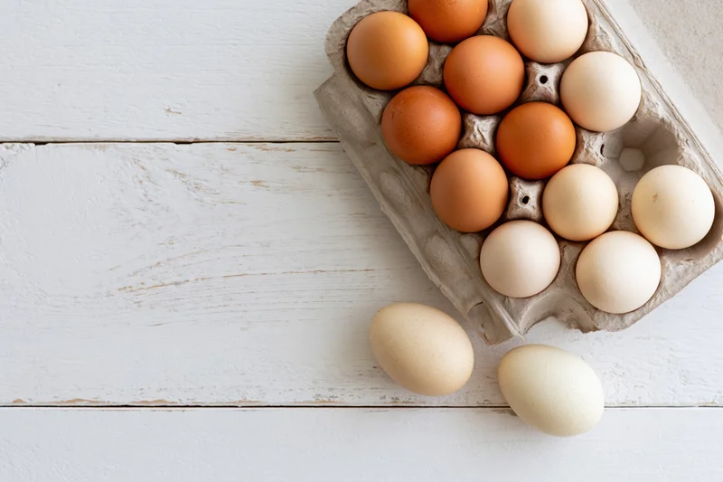 Farm-fresh chicken eggs in a grey cardboard carton arranged on a white wooden surface with two loose eggs beside it