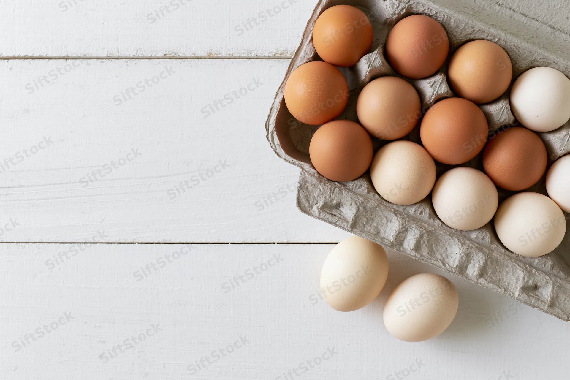 Dozen farm-fresh chicken eggs in a grey cardboard carton on a white wooden surface with two eggs spilled beside it