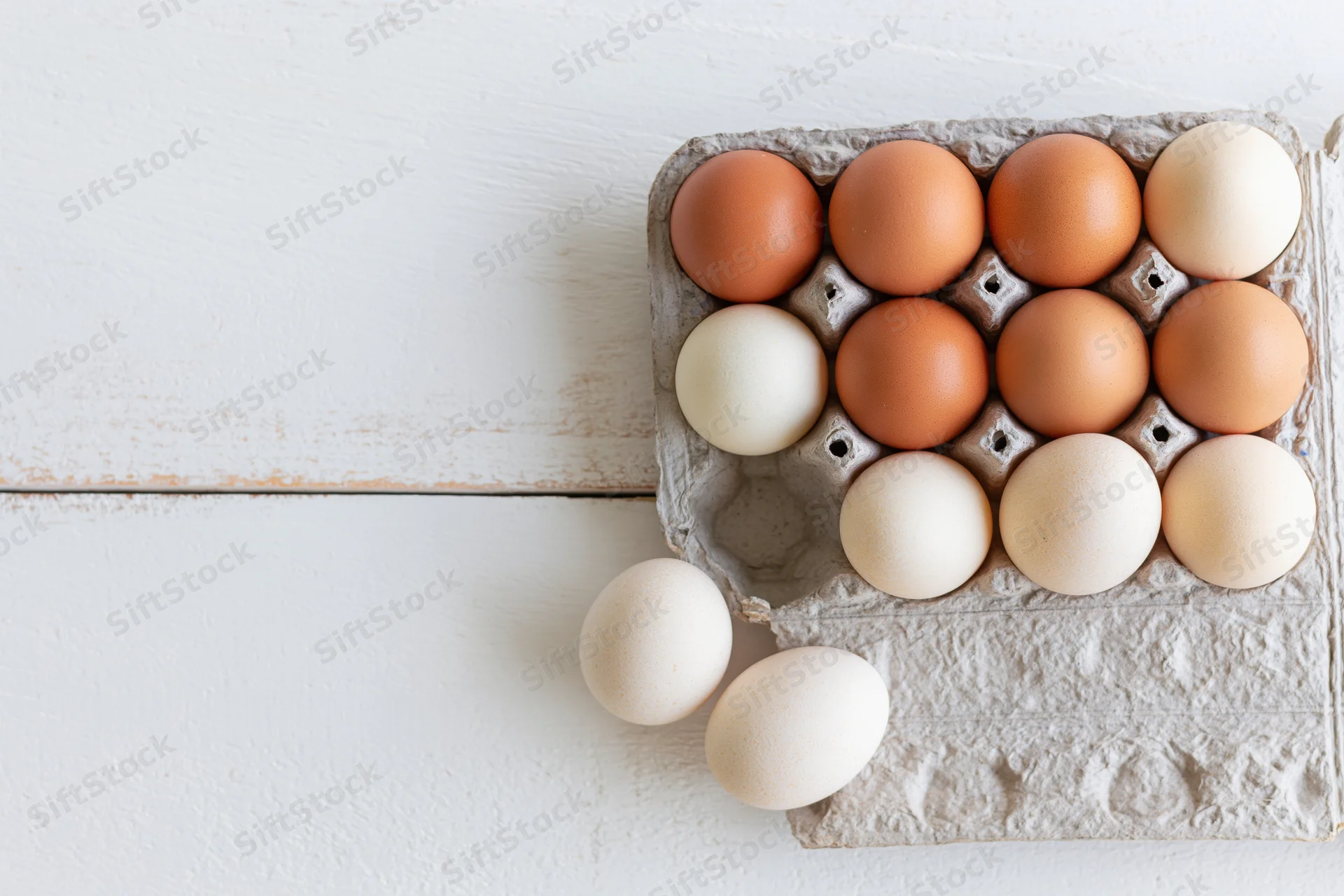 Dozen farm-fresh chicken eggs in a grey cardboard carton on a white wooden surface, some spilled beside it