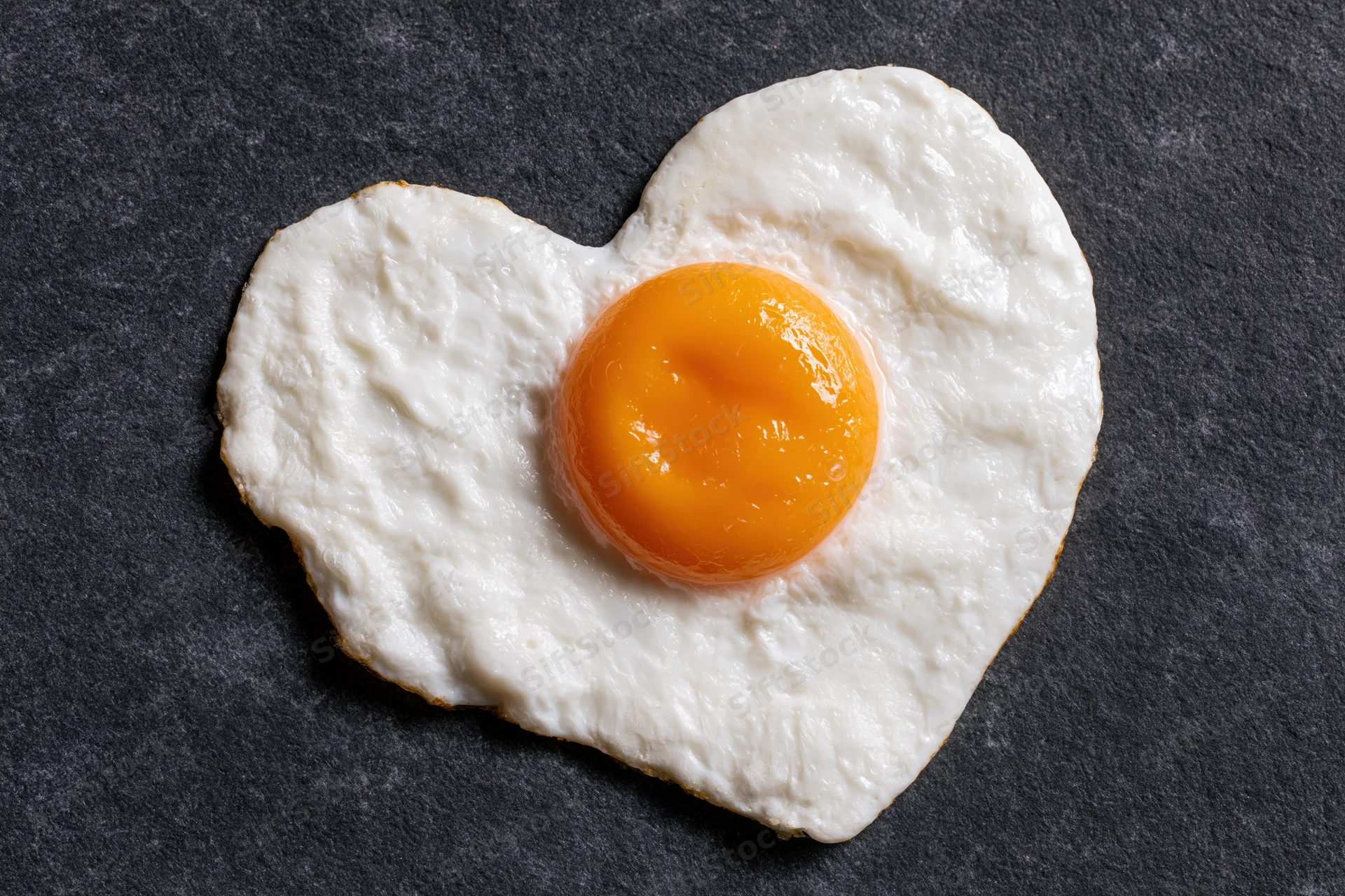 Fried egg cooked into heart shape sunny side up on dark slate surface