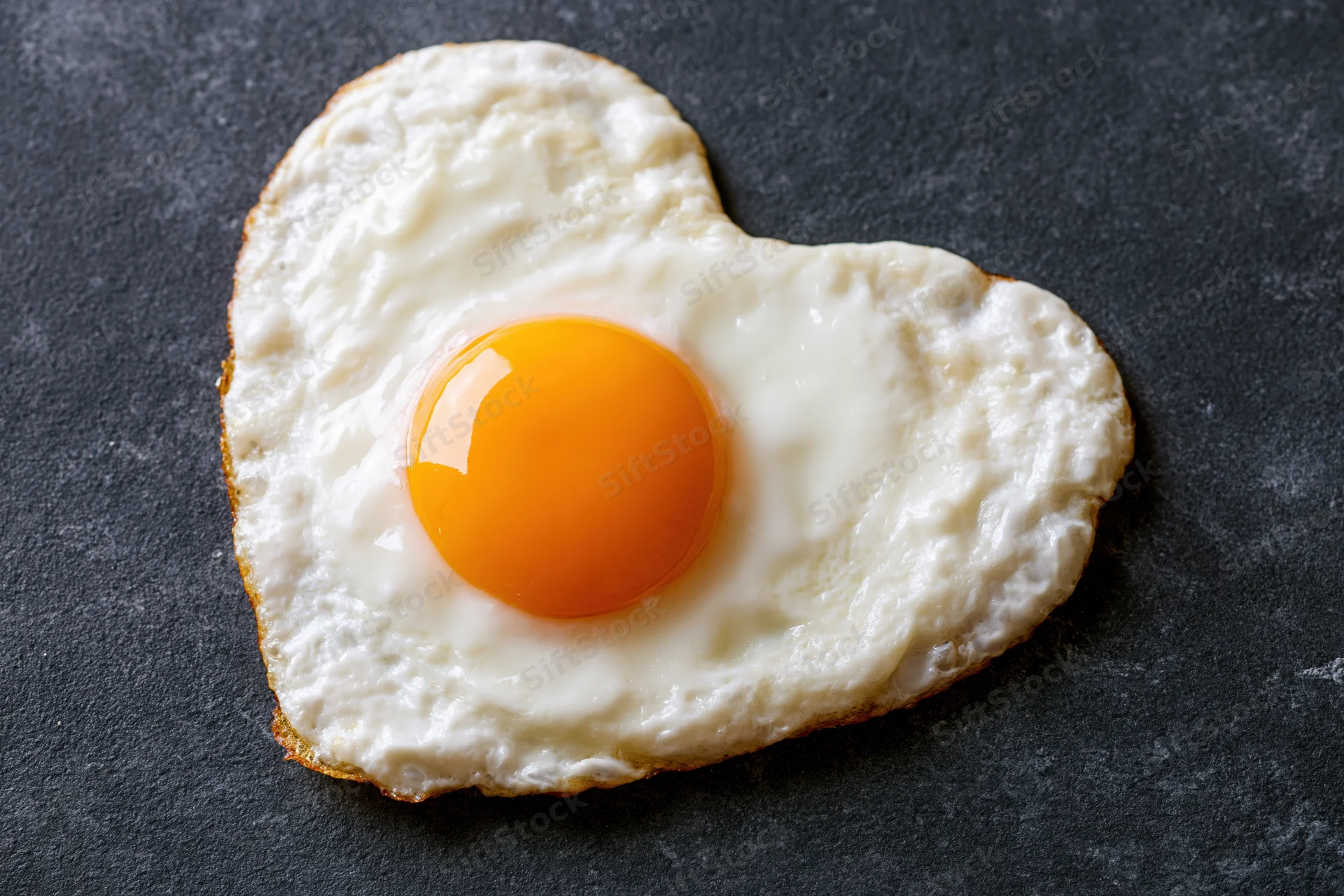 Fried egg cooked into heart shape sunny side up on dark surface