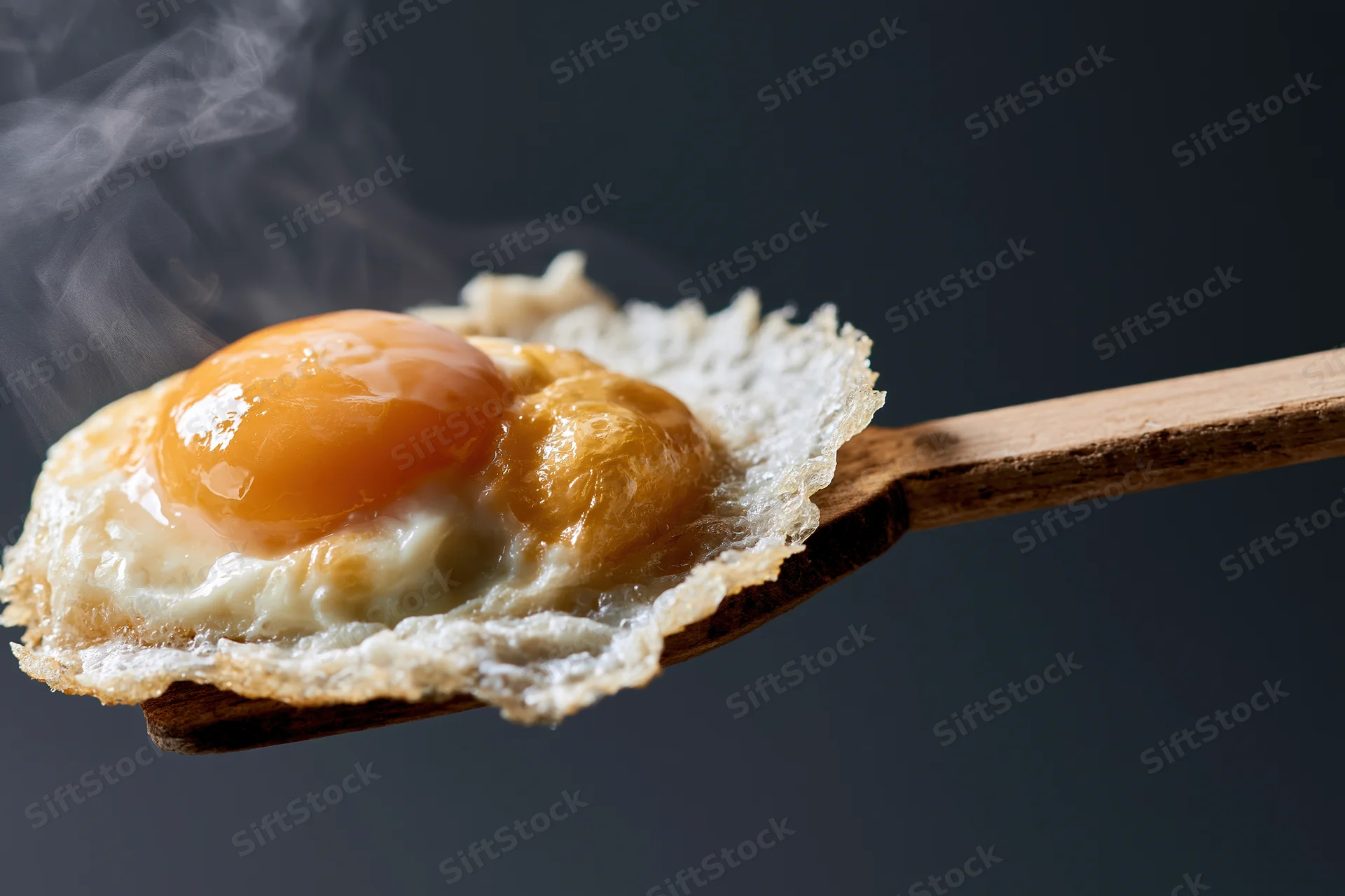 Fried sunny-side-up egg resting on a worn wooden spatula with steam rising against dark background