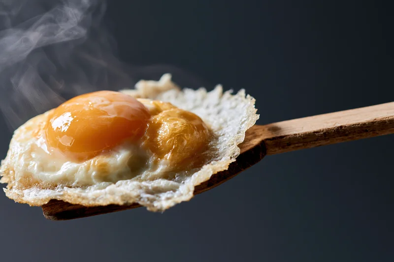 Fried sunny-side-up egg resting on a worn wooden spatula with steam rising against dark background
