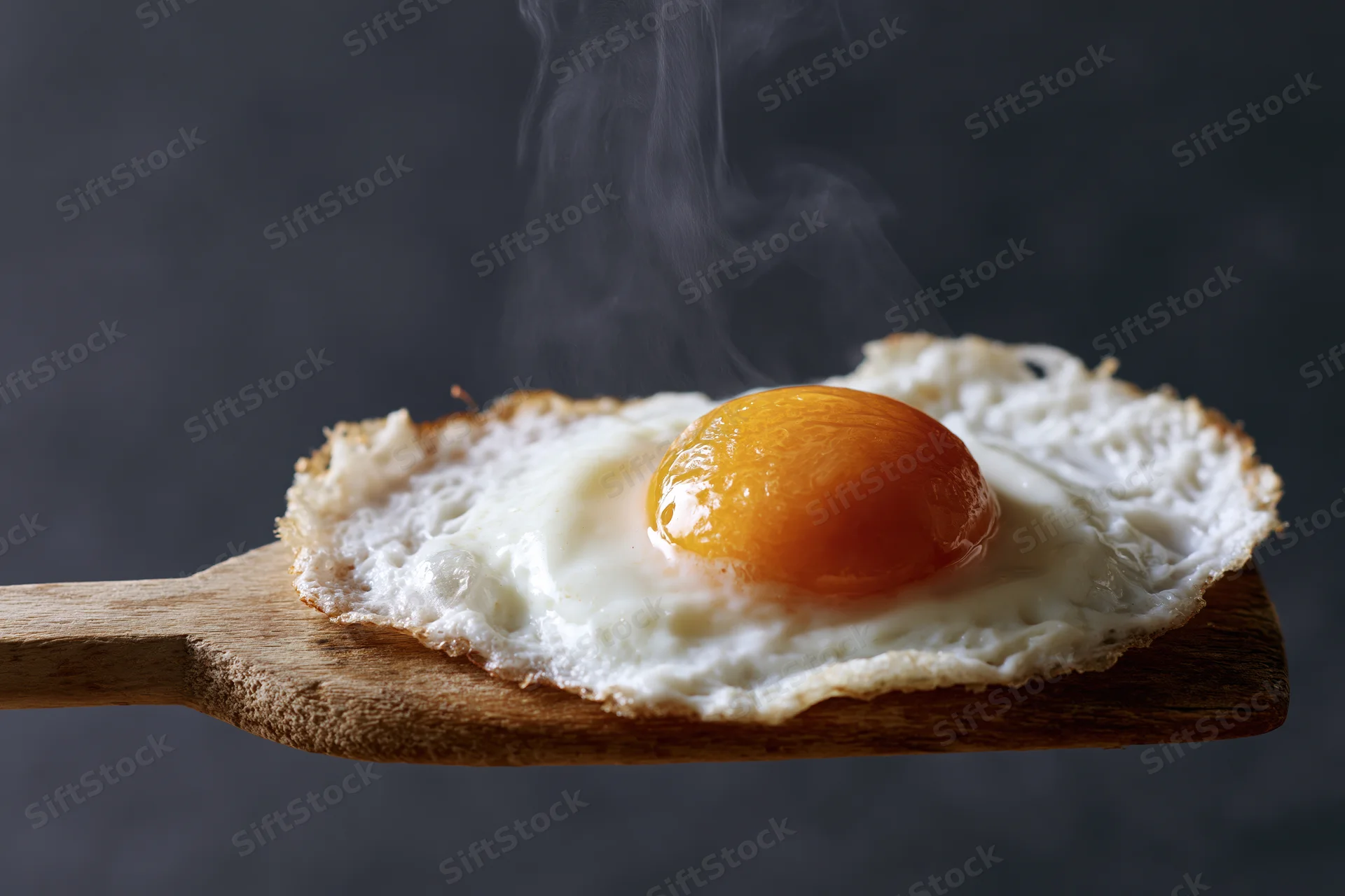 Fried sunny-side-up egg with steaming yolk resting on a worn wooden spatula against dark background