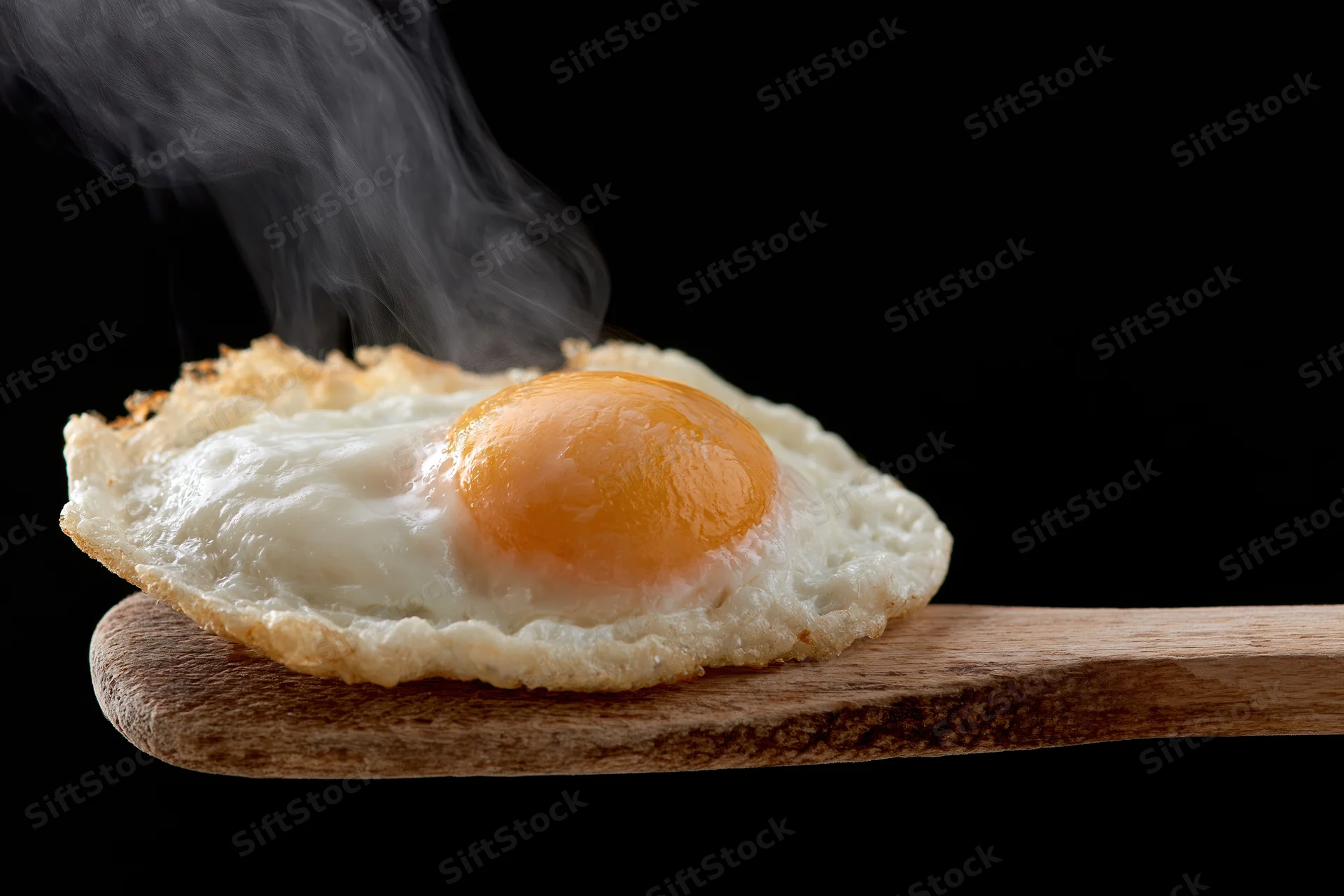 Fried sunny-side-up egg with steam rising on a worn wooden spatula against black background
