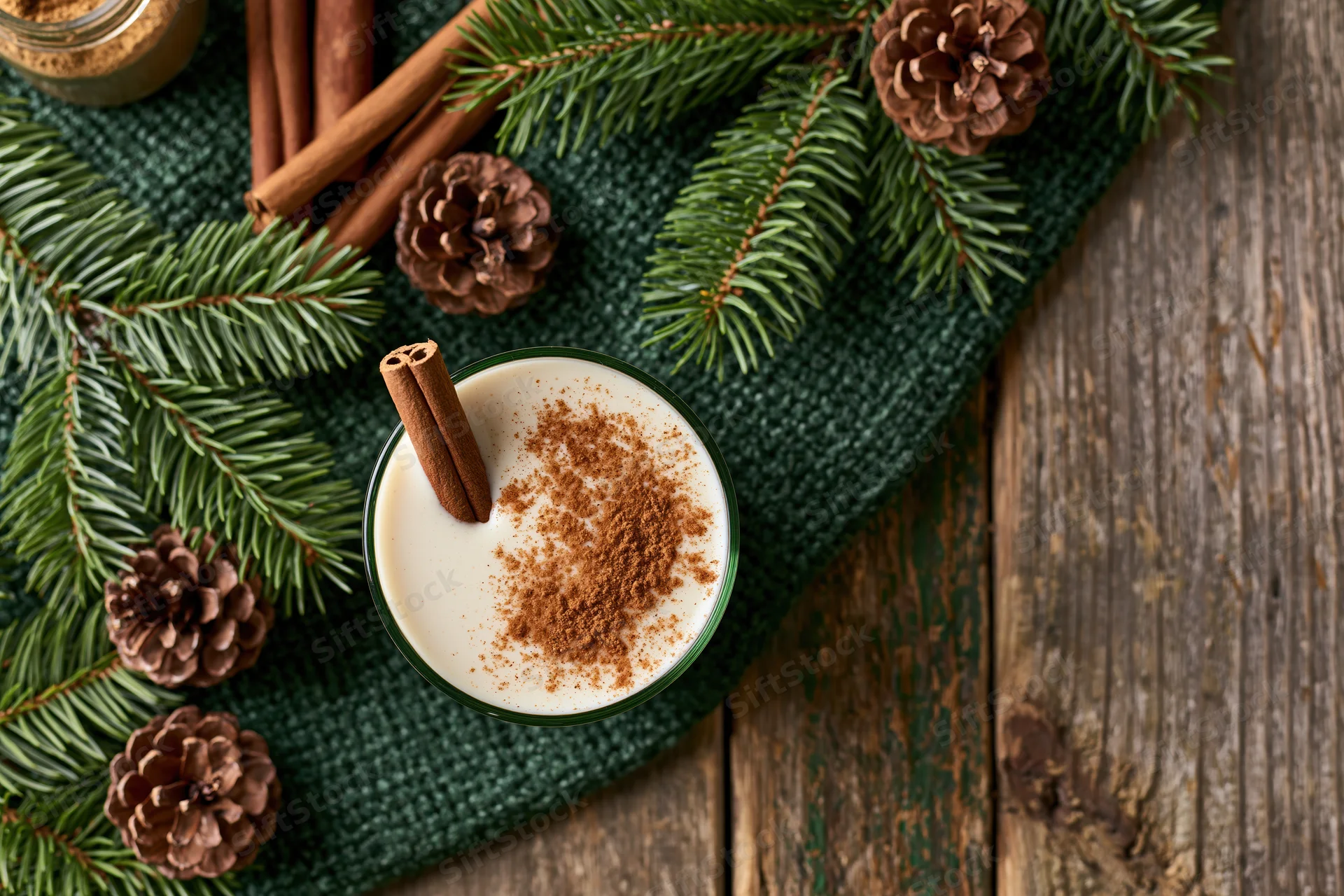Creamy eggnog topped with ground cinnamon and a cinnamon stick on a green cloth with pine branches and pinecones on a wooden table