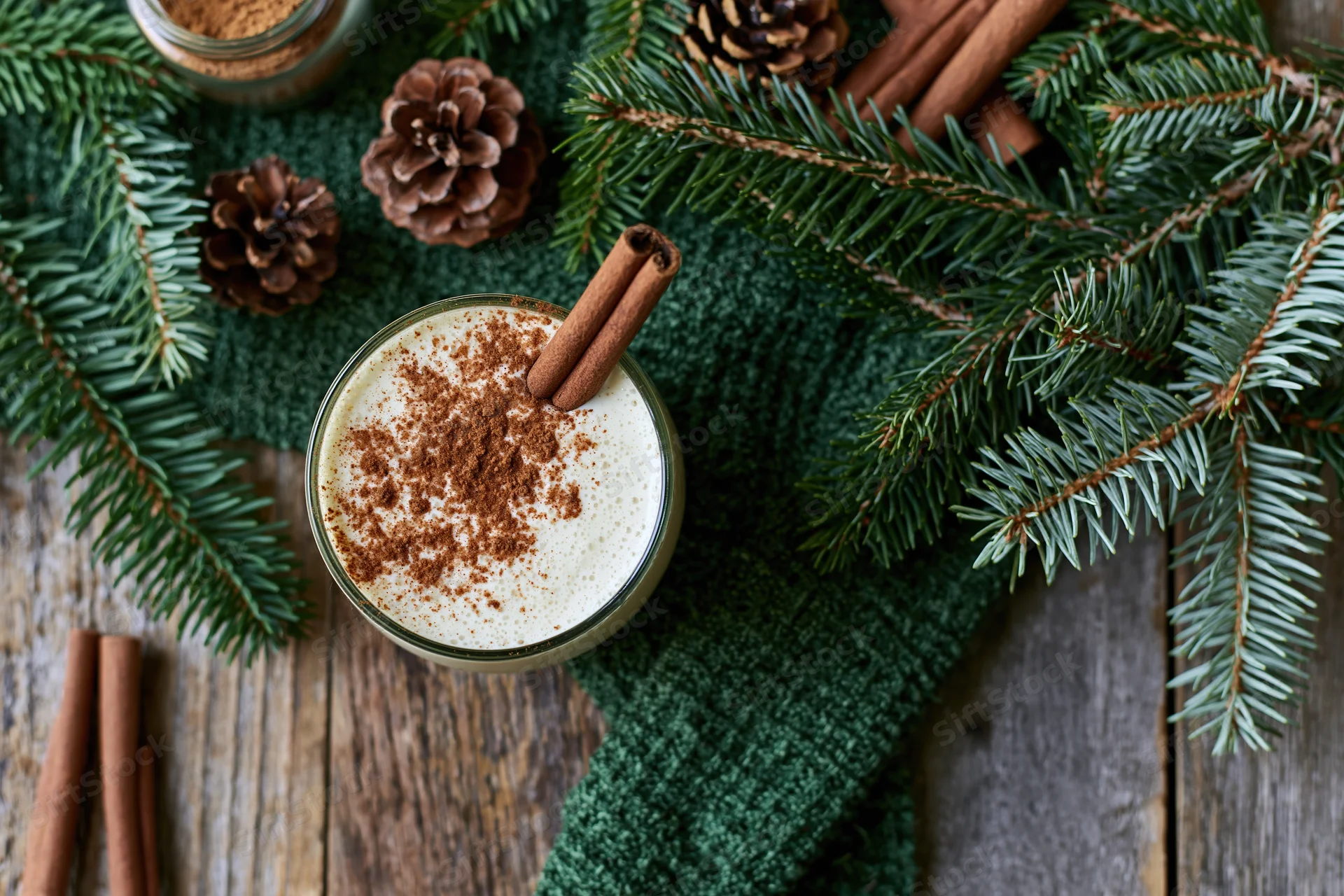 Creamy eggnog in a glass topped with cinnamon and a cinnamon stick on a wooden table with pine branches and pinecones