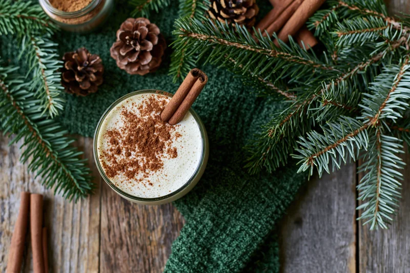 Creamy eggnog in a glass topped with cinnamon and a cinnamon stick on a wooden table with pine branches and pinecones