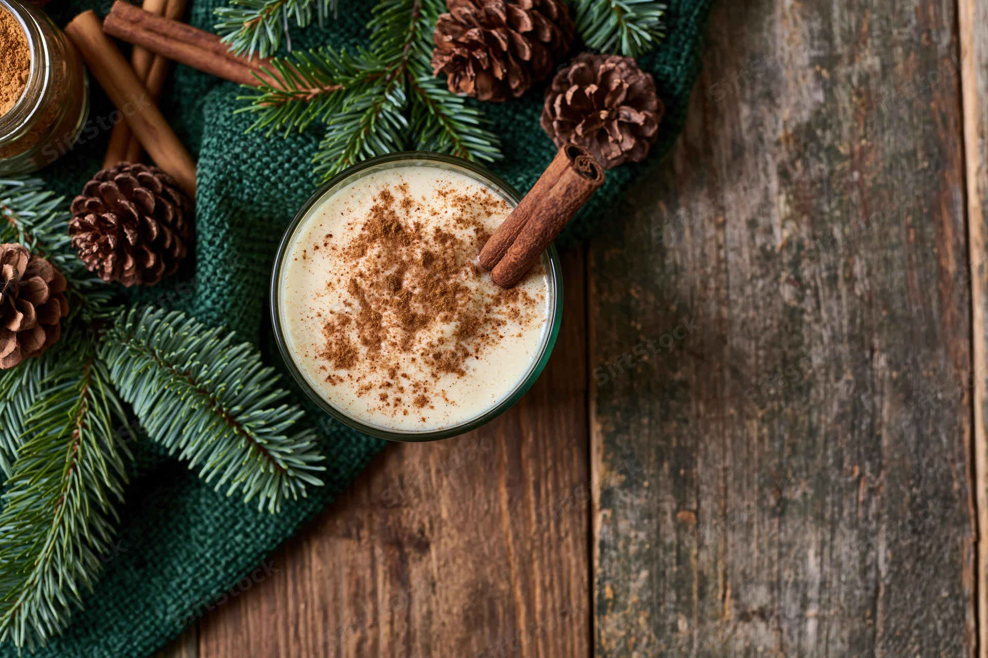 Creamy eggnog in a glass topped with cinnamon and a cinnamon stick on a wooden table with pine branches and pinecones