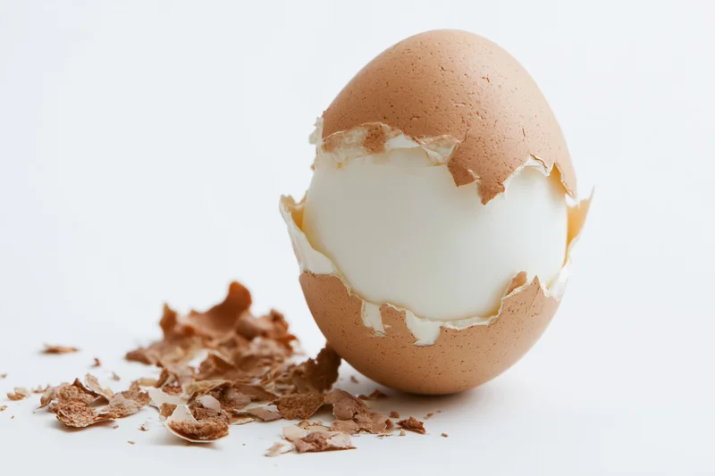 Hard-boiled egg with partially peeled shell on white surface, shell fragments scattered beside it