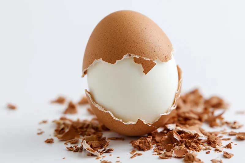 Hard-boiled egg with partially peeled shell resting on scattered shell fragments on white surface