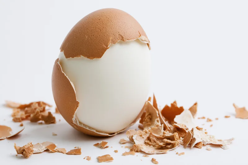 Hard-boiled egg with partially peeled shell on white surface surrounded by shell fragments