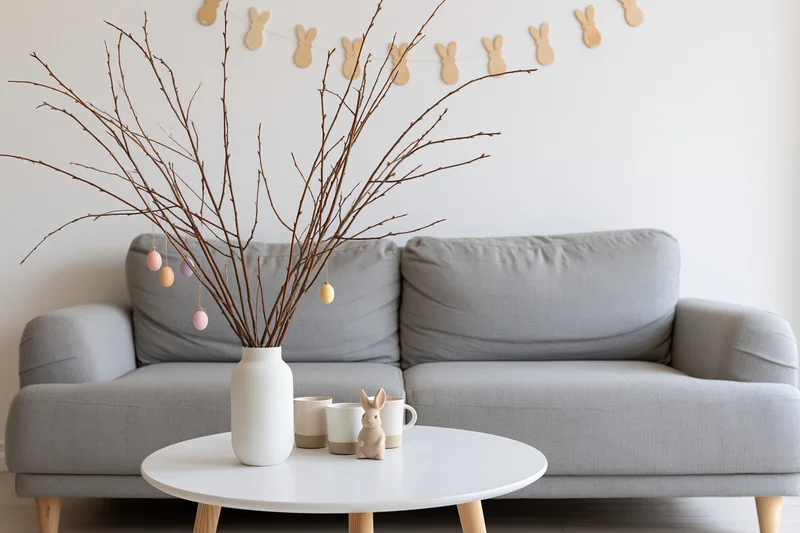 Easter decoration with bare branches and pastel eggs in a white vase on a coffee table in front of a gray sofa