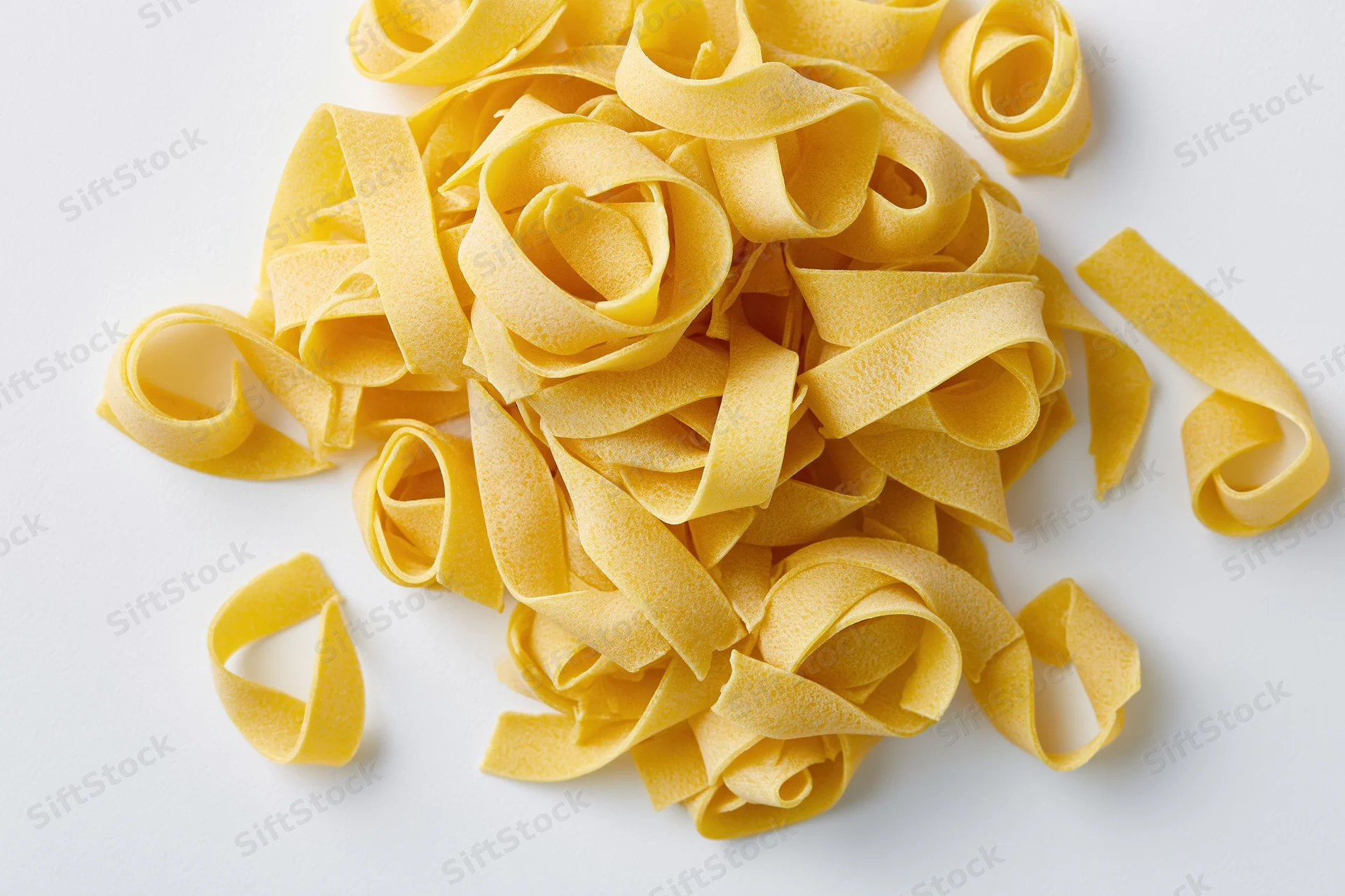Loose pile of dry uncooked egg pasta ribbons curled and scattered on white surface