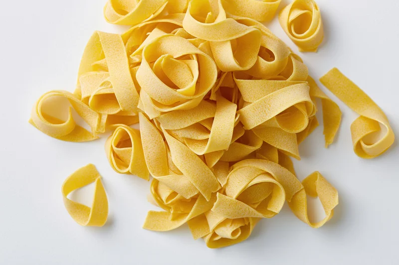 Loose pile of dry uncooked egg pasta ribbons curled and scattered on white surface