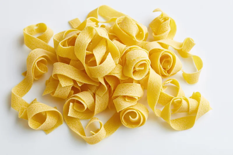 Dry uncooked egg pasta ribbons curled and piled on white surface