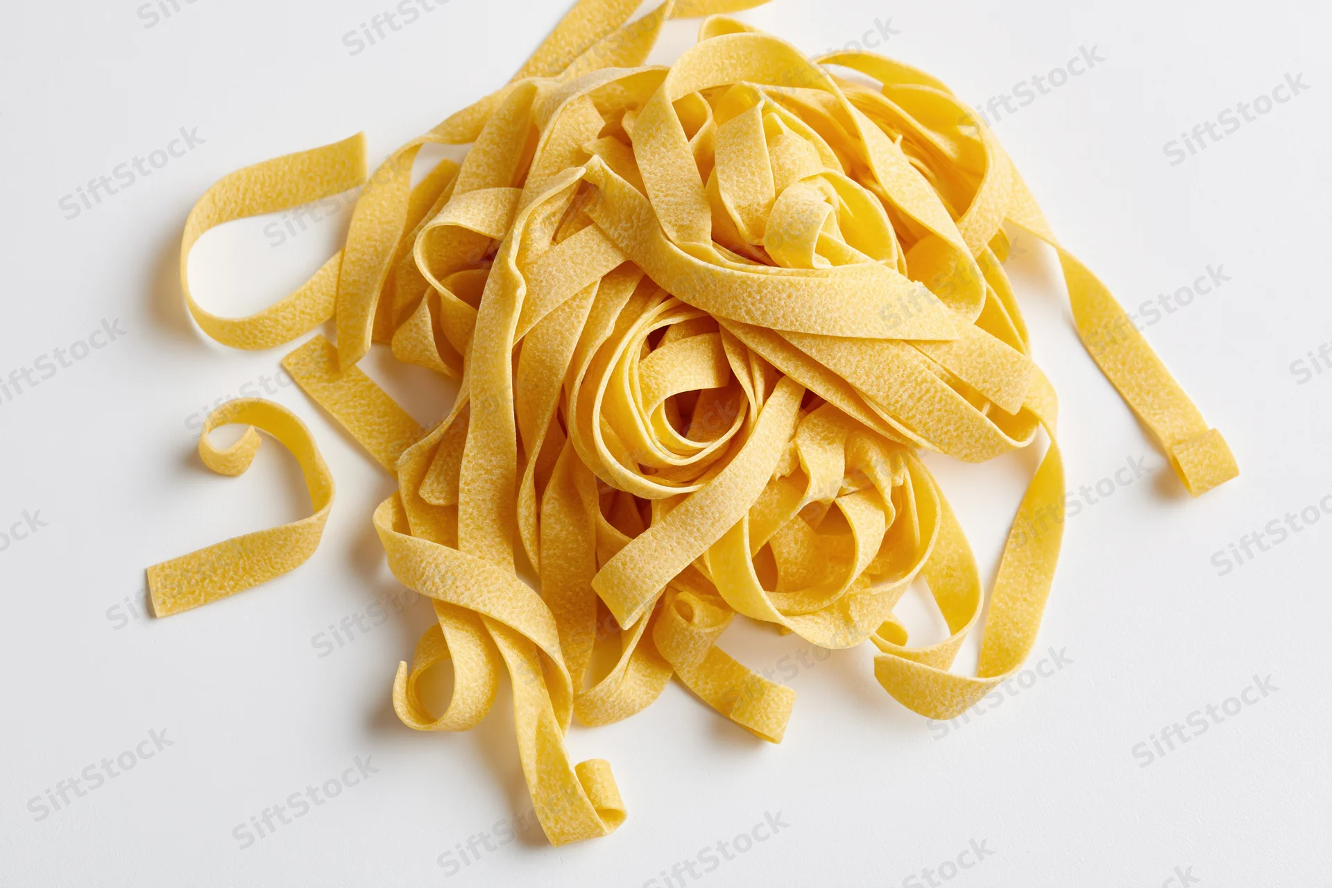 Loose pile of dry uncooked egg pasta ribbons curled and twisted on white background