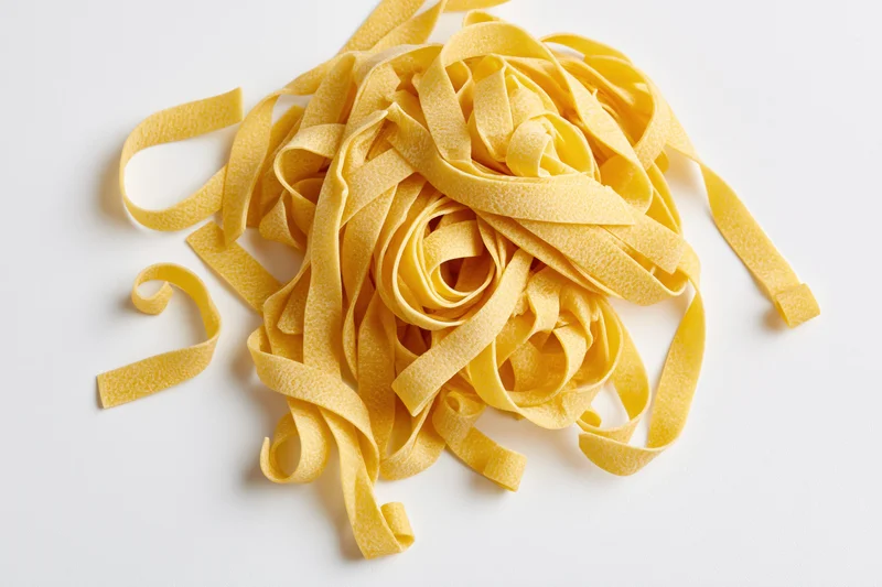 Loose pile of dry uncooked egg pasta ribbons curled and twisted on white background