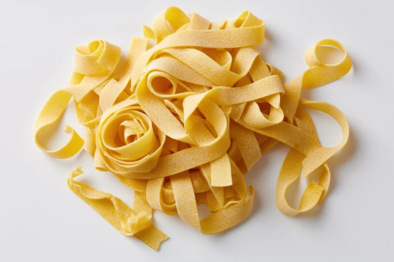 Loose pile of dry uncooked egg pasta ribbons curled and twisted on white background