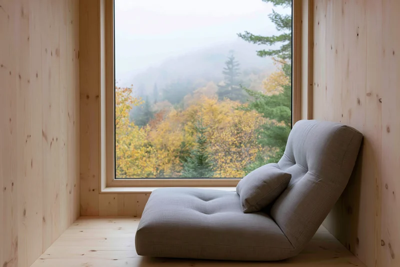 Gray chaise lounge in a pine-clad cabin reading nook facing autumn forest view through large window