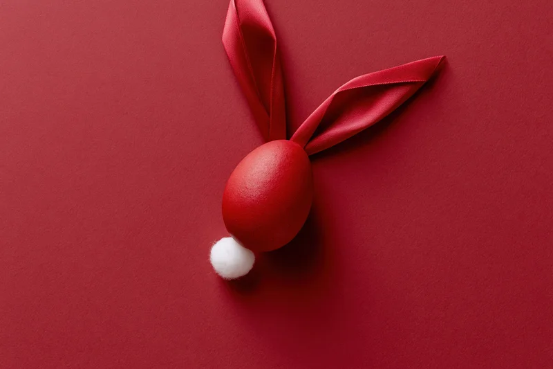 Red-dyed easter egg styled as bunny face with ribbon ears and white pompon tail on red background