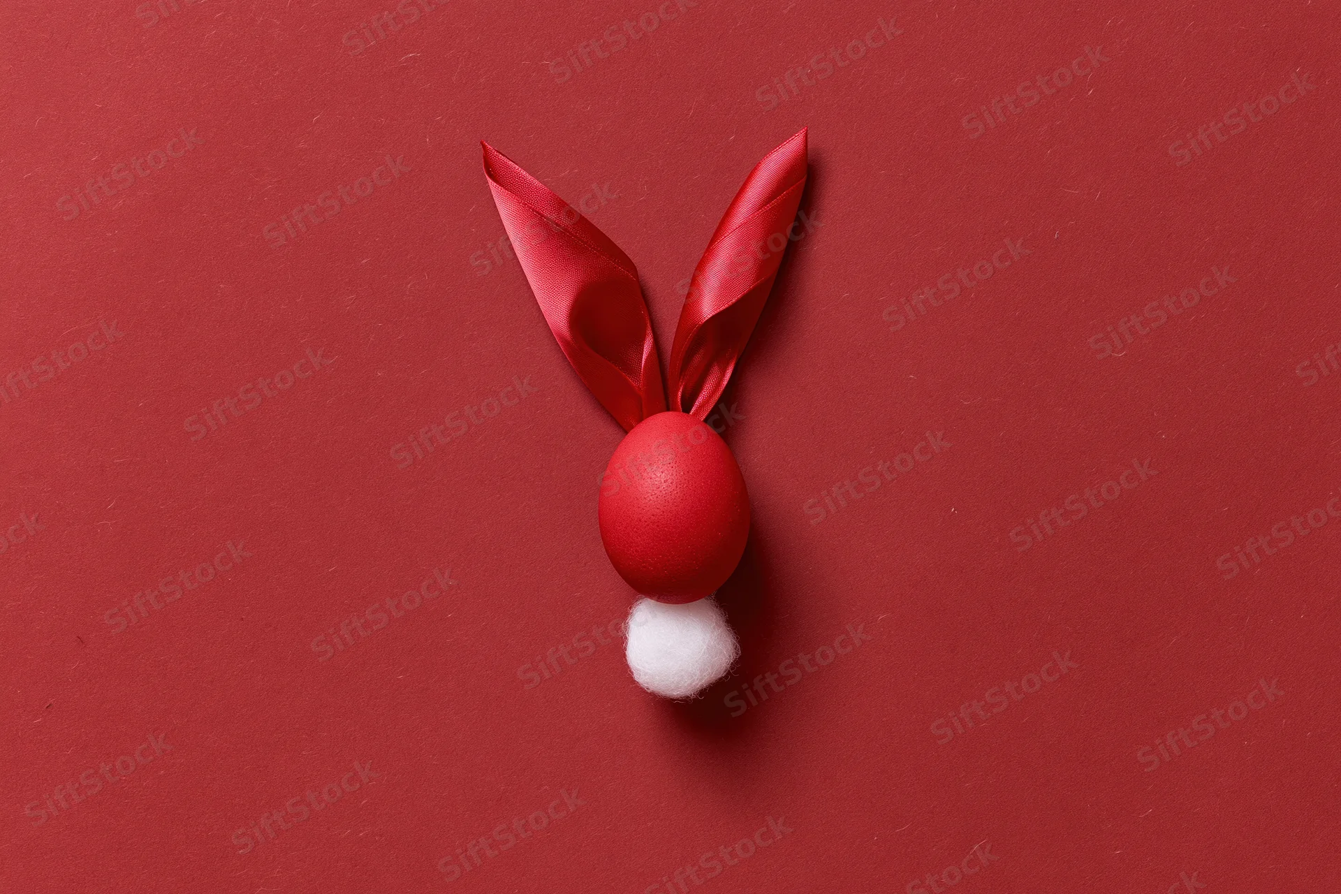 Red-dyed easter egg styled as bunny face with ribbon ears and cotton tail on red background