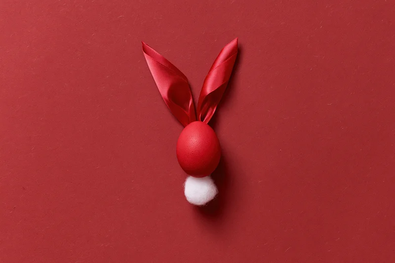 Red-dyed easter egg styled as bunny face with ribbon ears and cotton tail on red background