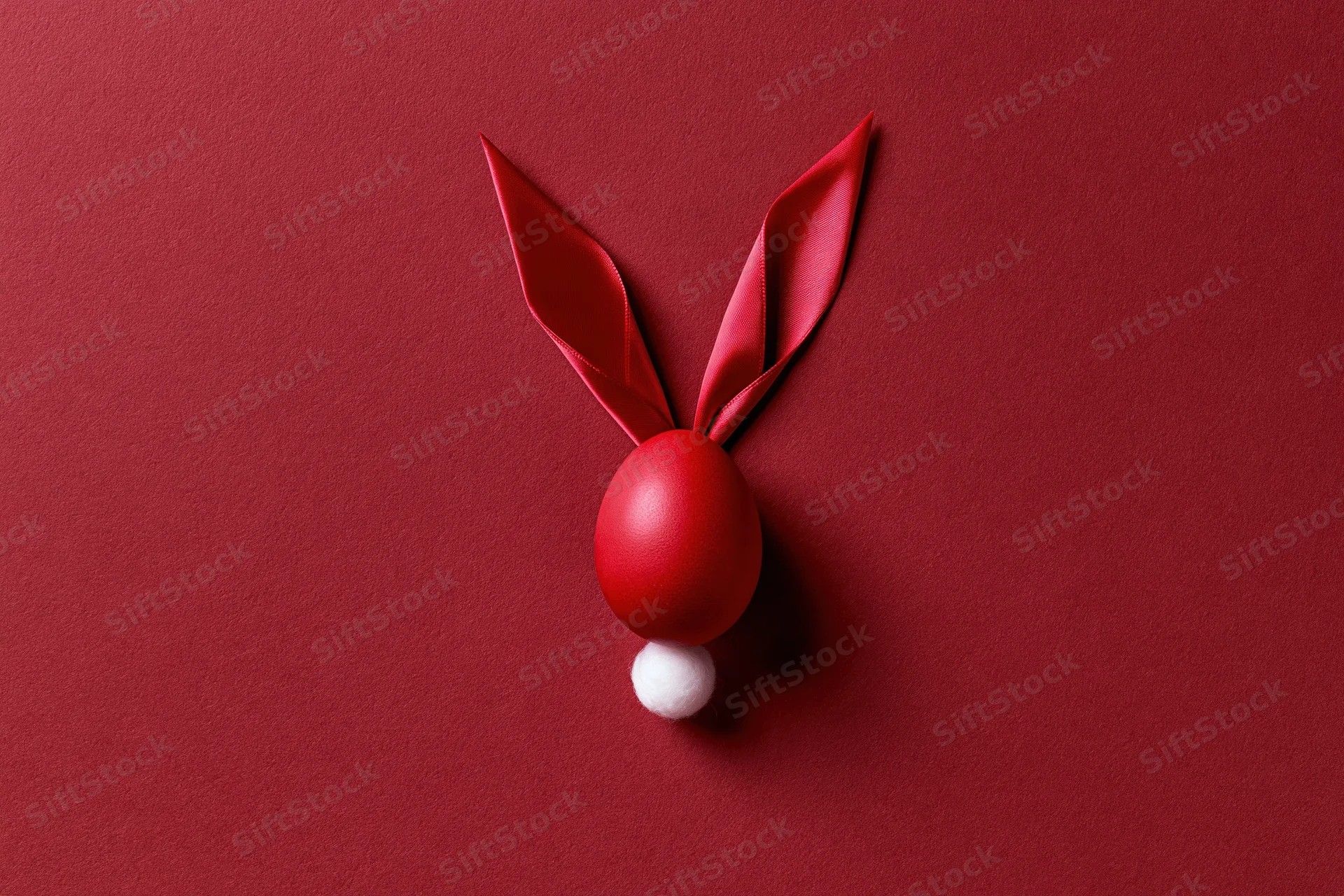 Red-dyed easter egg styled as bunny face with ribbon ears and white pompon tail on red surface