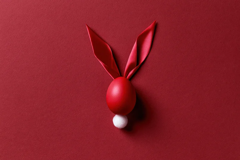 Red-dyed easter egg styled as bunny face with ribbon ears and white pompon tail on red surface