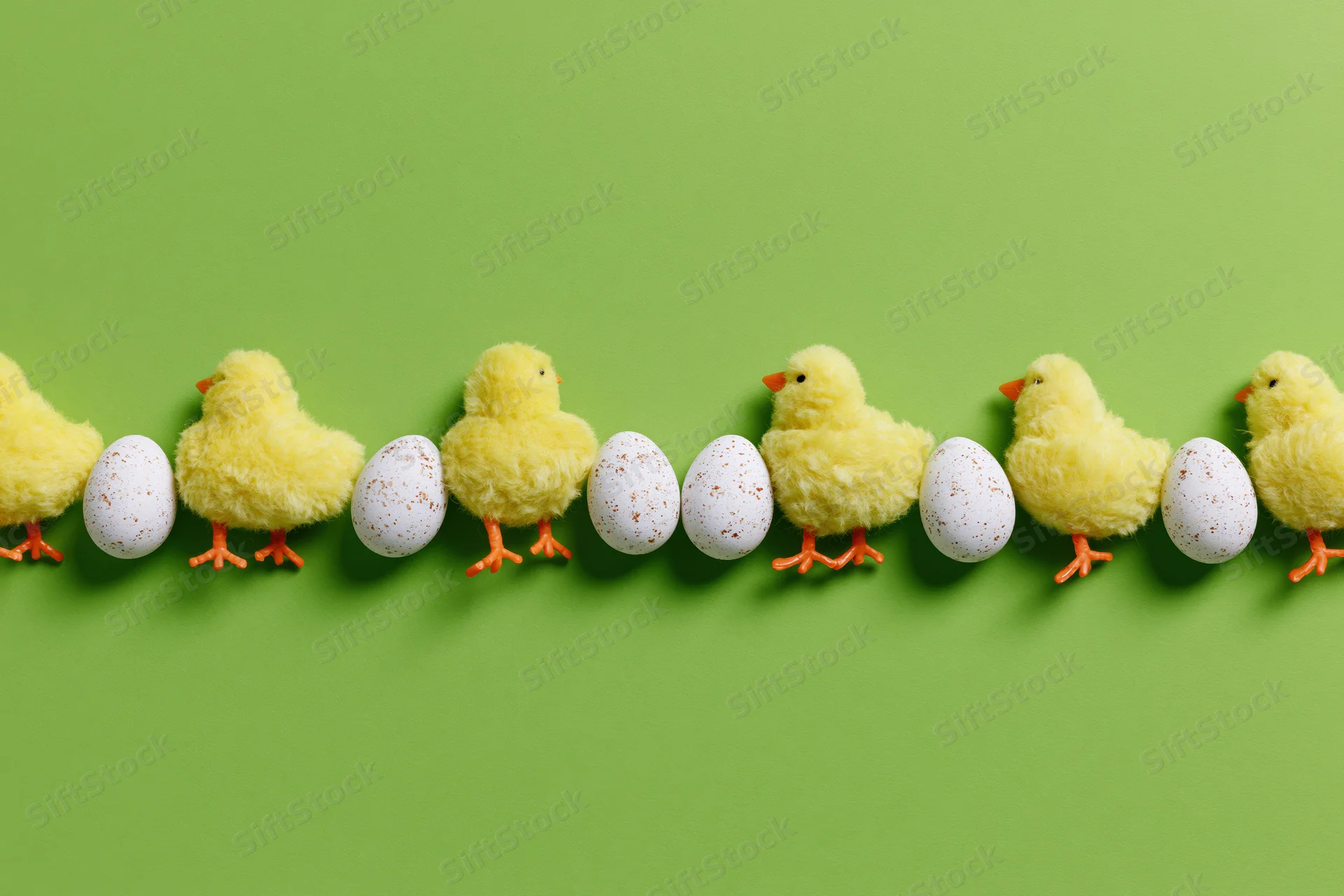 Row of fluffy yellow toy chicks and white speckled eggs arranged in a line on a green background