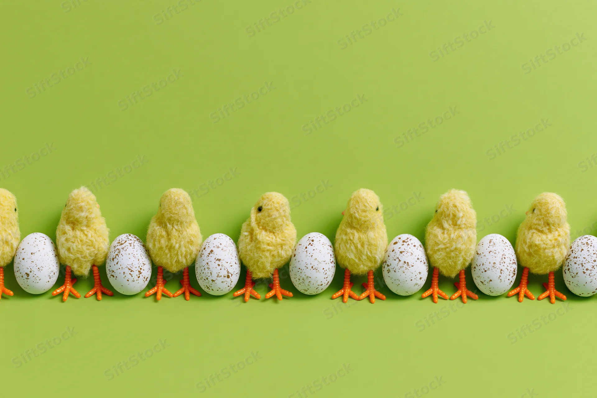 Row of fluffy yellow toy chicks with orange legs and speckled eggs on green background