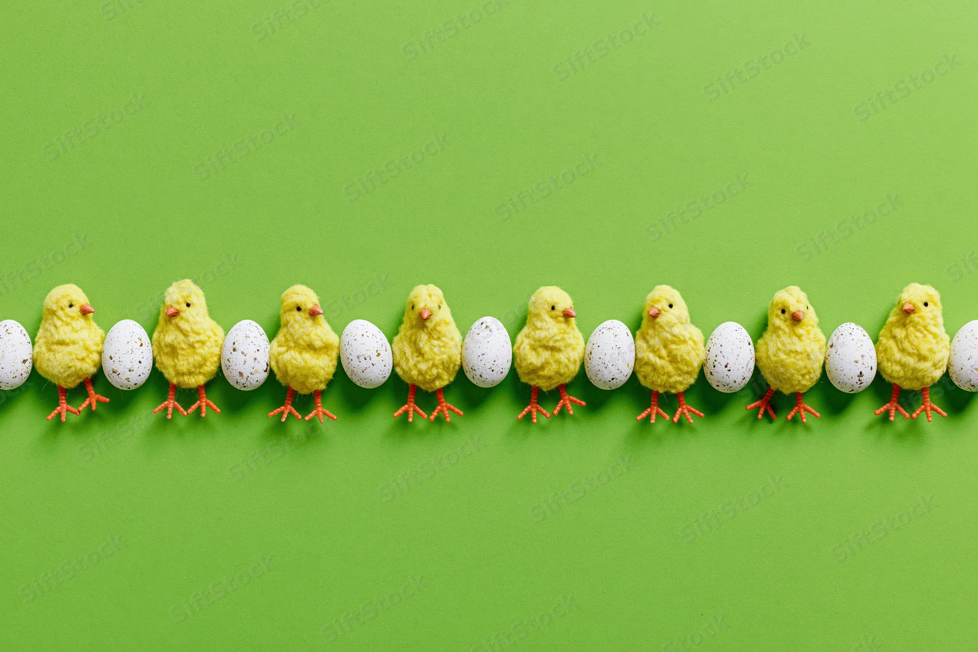 Row of yellow toy chicks with orange legs and white speckled eggs arranged in a line on green background