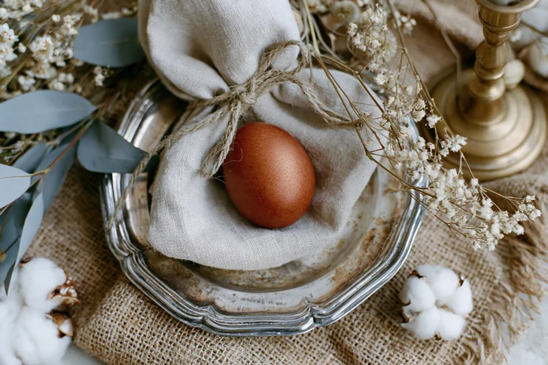 Terracotta-dyed easter egg on linen napkin with twine, arranged on silver plate with dried flowers and cotton, rustic flat lay on burlap surface