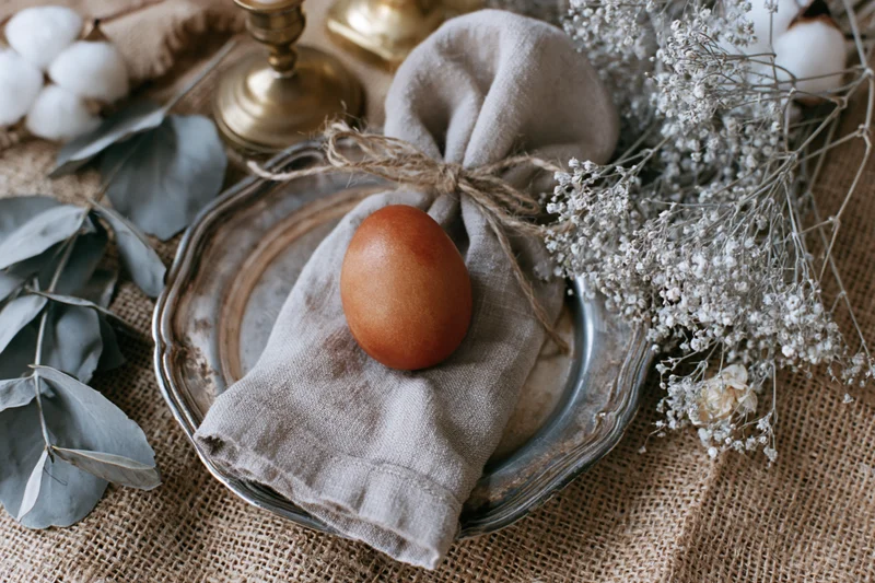 Terracotta-dyed easter egg on a linen napkin tied with twine, placed on a silver plate with dried flowers and eucalyptus on a burlap tablecloth